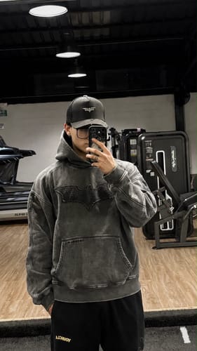 Customer's Batman Stealth Hoodie in a washed grey finish, being worn while taking a mirror selfie in a gym.