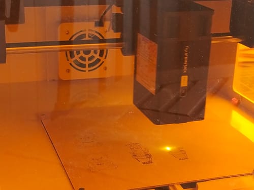 Customer's AtomStack P1 laser engraver active, etching cactus designs onto a metallic surface behind amber glass.