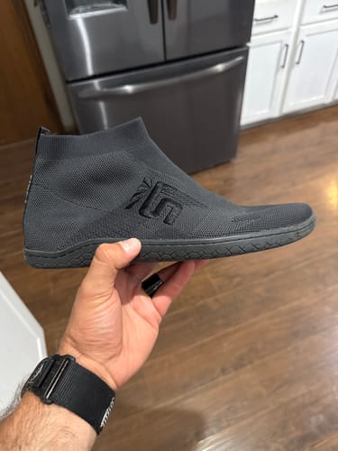 Reviewer holding the black Otishi 2.0 shoe, highlighting the sleek knit design and flat sole.
