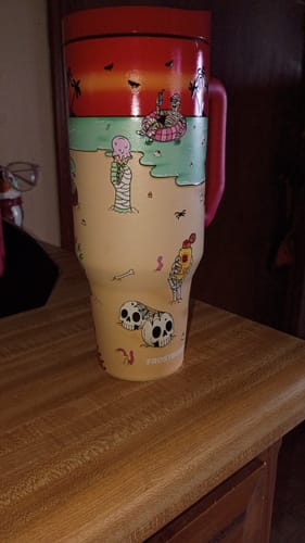 Customer's Thicc Buddy 40oz mug with a beach and skeleton graphic on a wooden table.