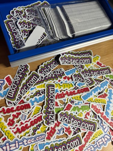 Customer's numerous custom vinyl stickers with vizi-tec.com branding, scattered on a table and inside a blue tray.