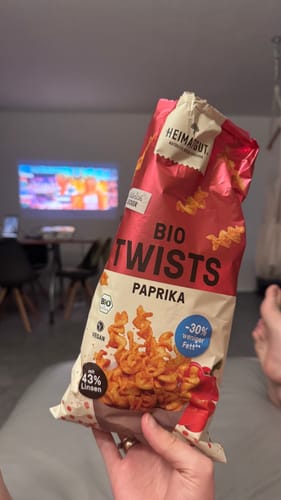 Customer's hand holding a bag of Bio Twists Paprika from the Heimatgut Probierbox inside a room.