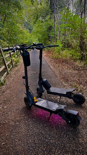 Customer's two Ausom L1 electric scooters parked on a paved forest path with pink underglow lights.