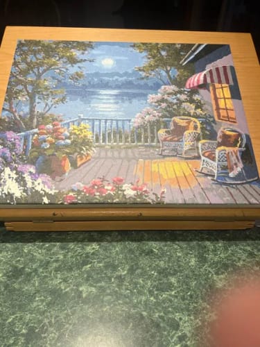 Reviewer's finished Custom Paint By Numbers landscape on an easel, depicting a porch with rocking chairs by the water.