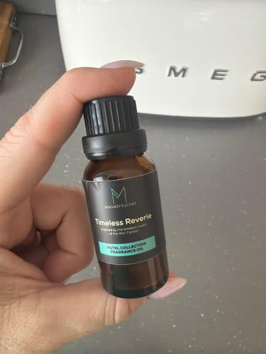 Reviewer's hand holding a brown bottle of Timeless Reverie fragrance oil in a kitchen setting.