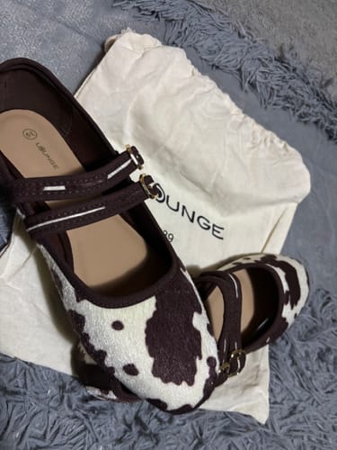 Customer's Zapato Ballerina Vaca Doble Hebilla con estampado de manchas marrones y blancas sobre una bolsa de tela.
