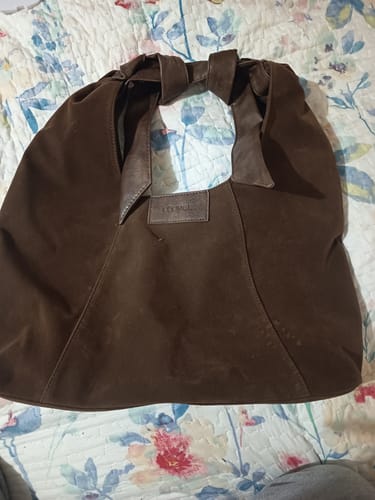 Reviewer's Cartera De Hombro Medialuna Nudo Suede Woman en color café, vista frontal sobre una superficie floreada.