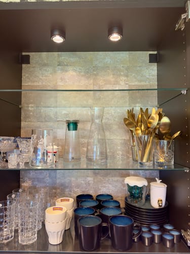 Customer's Rusty Gold kontaktplast applied as a backsplash inside an open, dark cabinet holding glassware and utensils.