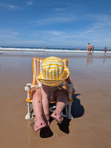 Customer's Brighton baby chair in yellow and white stripes, placed on the sandy beach with a child sitting inside.