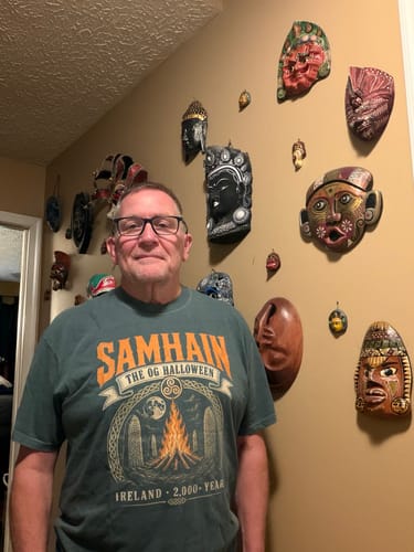 Customer's Samhain Og T-shirt featuring a green design with bonfire imagery, worn indoors in front of wall masks.