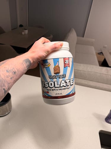 Customer's hand holding a white tub of UGLYFACE Isolate Protein V2.0 Birthday Cake flavor in a home setting.