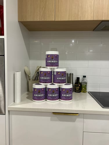 Customer's pyramid stack of six UGLYFACE AMINOS+ purple containers on a white kitchen counter.