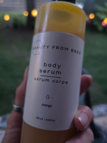Customer's hand holding a bottle of Beauty From Bees Body Serum, orange scent, with a blurred outdoor background.