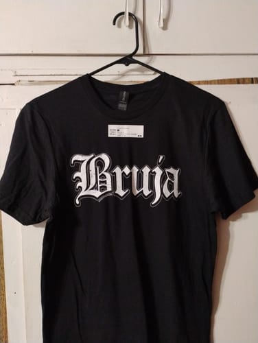 Reviewer's black Bruja T-shirt hanging on a plastic hanger against a white door.