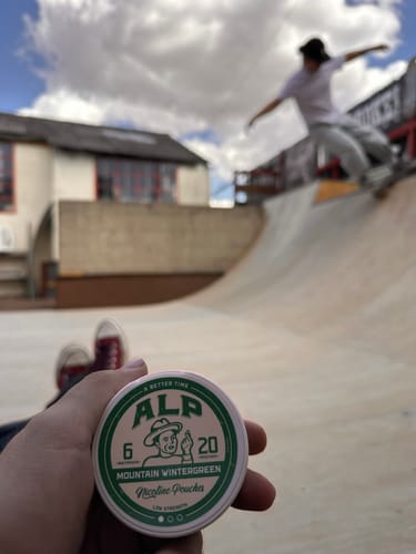 Customer's hand holding a round, white and green ALP Mountain Wintergreen tin at a skatepark.