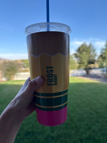 Customer's hand holding a pencil-themed To-Go Buddy tumbler with a blue straw against a bright, grassy outdoor background.