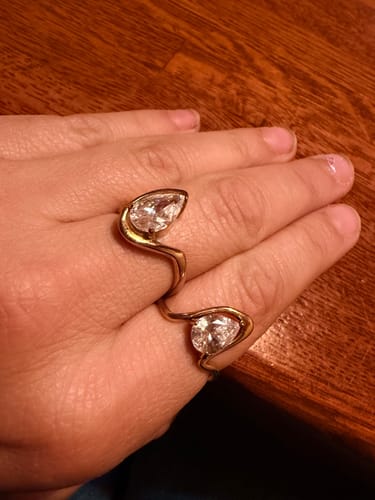 Customer's hand wearing two gold-toned Navae Birthstone Rings, each featuring a clear, pear-cut stone.