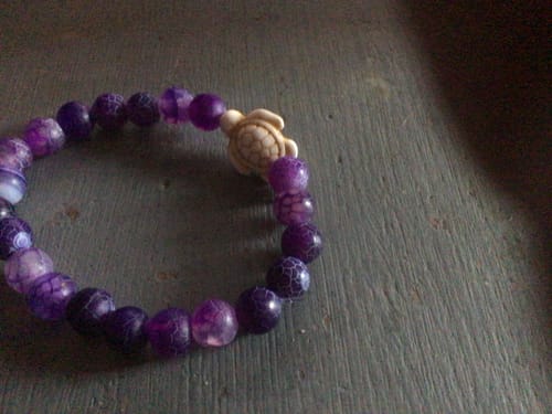 Customer's purple beaded Journey Bracelet with a turtle charm resting on a textured wooden surface.