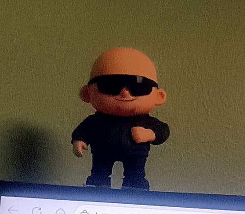 Customer's custom action figure, a bald figurine wearing black sunglasses and a black outfit, standing against a wall.