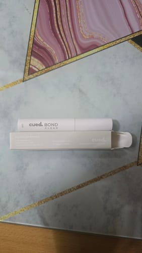 Kunde zeigt die weiße BOND Wimpernkleber-Verpackung und den Applikator auf einer strukturierten Oberfläche.