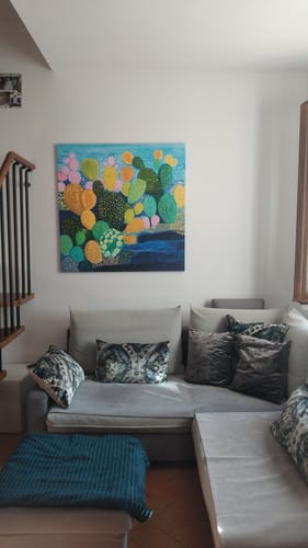 Customer's Oasi di Fichi painting with colorful cactus shapes displayed on a wall above a grey living room sofa.