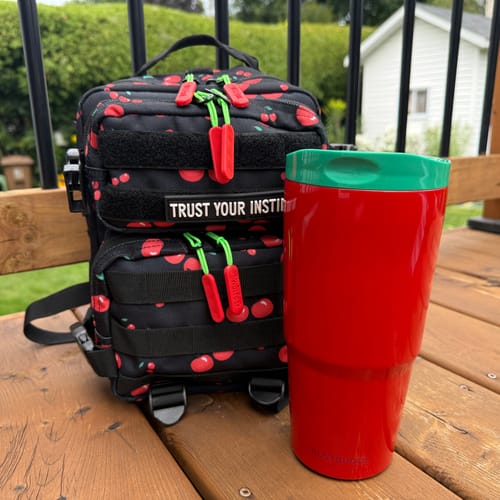 Customer's shiny red To-Go Buddy tumbler with a green lid, sitting on a wooden deck next to a black backpack.