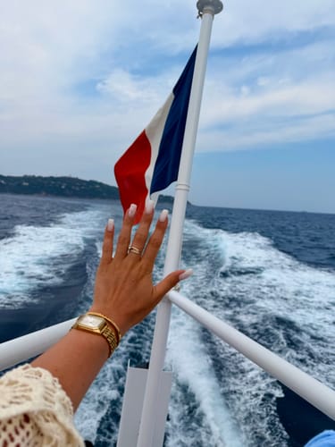 Reviewer's gold Ava Necklace and matching gold watch worn on a hand on a boat with a flag in the background.