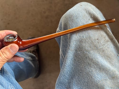 Customer's hand holding a long, brown glass pipe with a packed bowl against blue jeans.