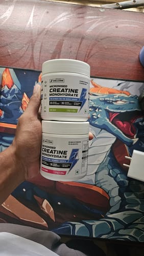 Reviewer's hand holding two stacked white tubs of Wellcore Micronised Creatine Monohydrate over a colorful desk mat.