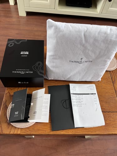 Customer's 50ml Fragrance & Body Wash Bundle, including a black box, a soft dust bag, and paper documents on a table.