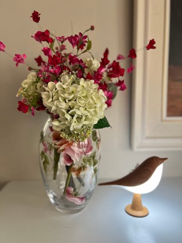 Customer's Glow Bird lamp glowing next to a floral vase on a table as a home decor addition.
