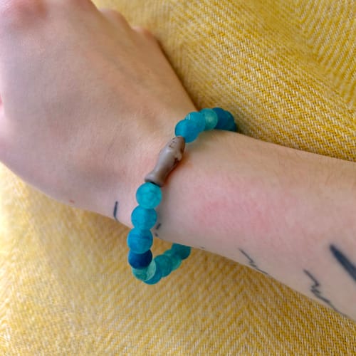 Customer's blue Voyage Bracelet with a small shark charm, worn on their wrist against a yellow textured background.