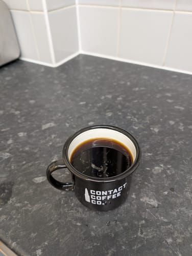 Customer's black enamel Contact Coffee mug filled with fresh coffee on a kitchen counter.