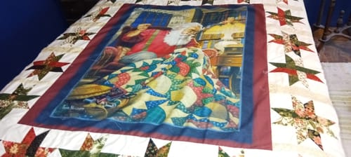 Reviewer's Merry Manor Jelly Roll fabric used as a patterned border around a Santa quilt panel on a bed.
