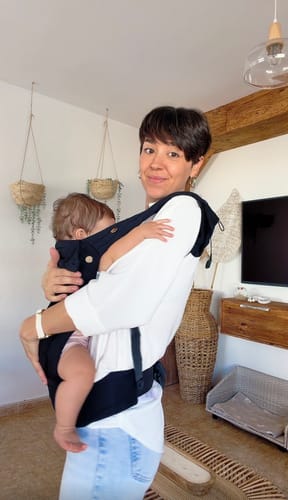 Customer's black HUG Ergonómica baby carrier worn by a person, carrying a baby in a neutral home setting.