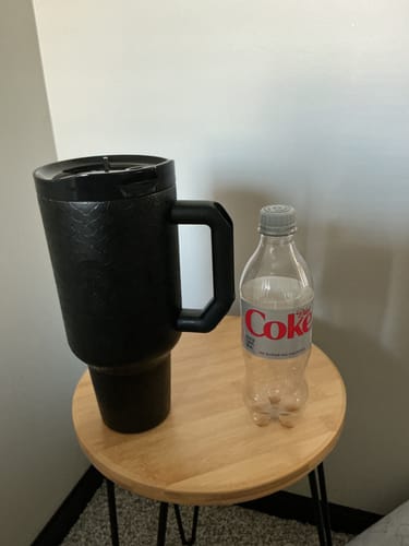 Customer's black Thicc Buddy 50oz mug placed on a small wooden side table next to a plastic water bottle.