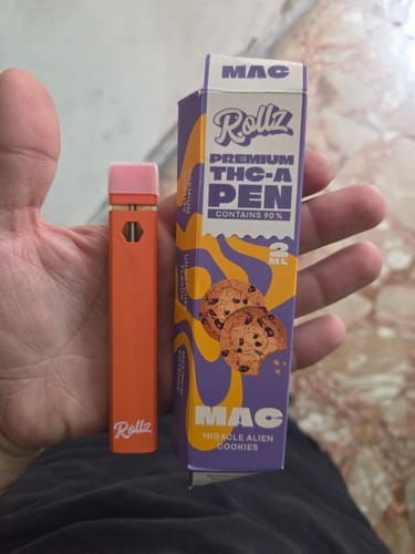 Customer's hand holding a bright orange THC-A Vape pen next to its purple MAC Miracle Alien Cookies packaging.