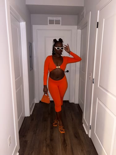 Customer's orange Naked Cutout Capri Jumpsuit with gold hardware, paired with orange heels in a hallway.