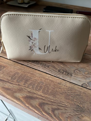 Kunden-Prio+ Kosmetiktasche in Beige mit floralem U-Initial und dem Namen Ulrike auf einem Holztisch.