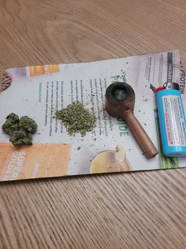 Customer's Godfather OG THCA Flower buds and ground cannabis on paper next to a wooden pipe and a light blue lighter.