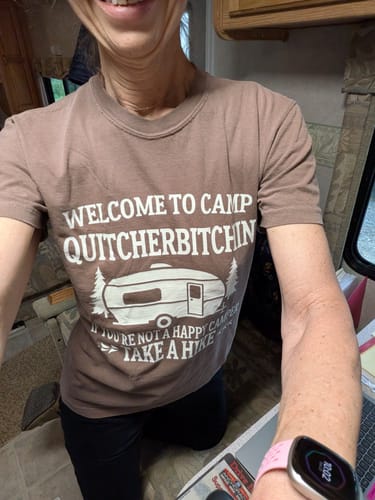 Customer's brown Camp Quit T-shirt with vintage camper graphic and text, worn in a casual indoor setting.