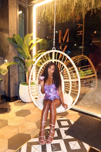 Customer's Sequin Open Back Mini Dress, sparkling in purple while sitting in a hanging chair.