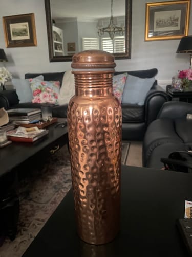 Customer's hand-hammered copper Ayurveda water bottle standing on a black table in a living room.