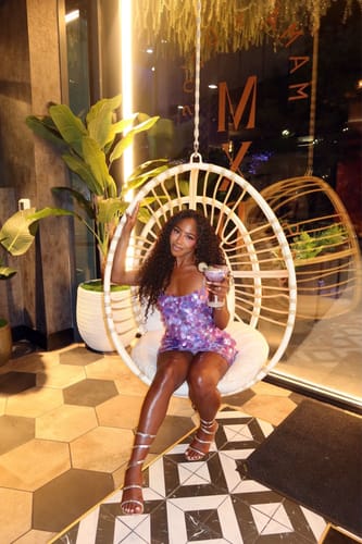 Customer's Sequin Open Back Mini Dress, sparkling in purple while sitting in a hanging chair.
