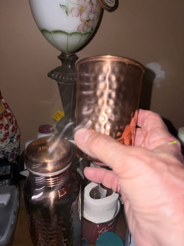 Customer's hand holding a hammered copper glass next to a copper water bottle, in a home setting.