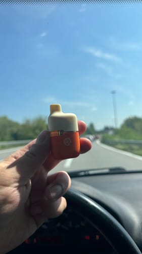 Reviewer's hand holding a compact, orange and cream colored THC-A vape device inside a car.