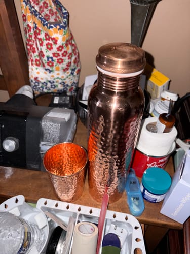 Customer's hand-hammered copper water bottle and cup from Certified Ayurveda sitting on a cluttered nightstand.