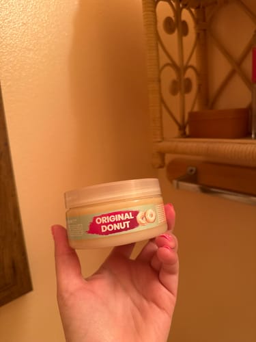 Reviewer's hand holding a jar of Body Glaze Original Donut cream in a room.