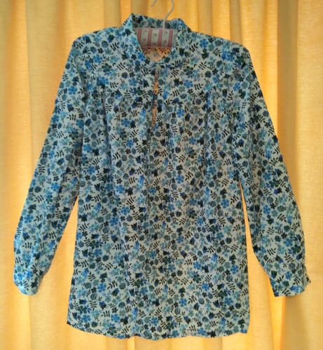 Customer's floral blouse sewn from cotton poplin fabric, hanging against a yellow curtain.
