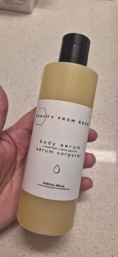 Customer's hand holding a bottle of unscented Beauty From Bees body serum against a bathroom counter.